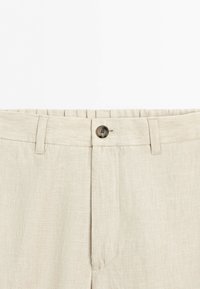 Beige linen trousers with belt loops, a button closure, and a visible waistband seam, shown from the waist to mid-thigh.