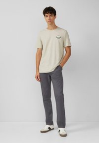 Lichtgrijs T-shirt met korte mouwen en een gestructureerde afwerking, met een donkergroene print. Combinatie met donkergrijze broek en witte sneakers.