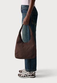 Borsa hobo in suede marrone con una fascia ampia, texture morbida, forma curvata e dettaglio di cucitura singola, tenuta da una persona che indossa jeans blu e scarpe da ginnastica.