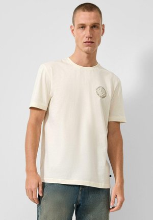 Man met kort haar die een crèmekleurig T-shirt draagt met een klein rond logo op de borst en vervaagde blauwe spijkerbroek, staand tegen een effen achtergrond.