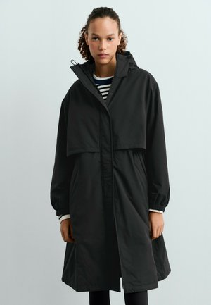 Femme aux cheveux bouclés portant un long manteau noir à capuche par-dessus une chemise rayée, debout devant un fond clair uni.
