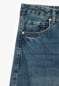 Jeans de mezclilla en un azul medio, con un bolsillo frontal, costuras doradas y una tela texturada con ligeros desgastes y detalles de desvanecimiento.