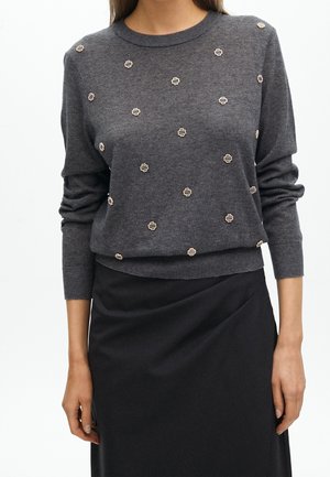 Frau trägt einen dunkelgrauen Langarm-Pullover mit kleinen weißen und schwarzen gestickten Punkten, kombiniert mit einem schwarzen Rock.