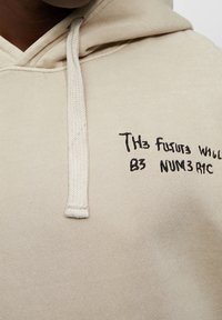 Beżowa bluza z kapturem z ściągaczem i czarnym haftem tekstu: "TH3 FUTUR3 WILL B3 NUM3RIC." Widoczna jest miękka faktura materiału.