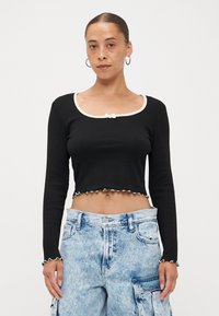 Black ribbed crop top with long sleeves, white scalloped trim, and a small bow detail at the neckline. Paired with light blue cargo pants.