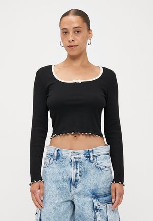 Black ribbed crop top with long sleeves, white scalloped trim, and a small bow detail at the neckline. Paired with light blue cargo pants.