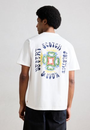 Man met afro kapsel draagt een wit T-shirt met kleurrijk abstract ontwerp en de tekst "Desert Scotch & Soda Solstice" op de achterkant.