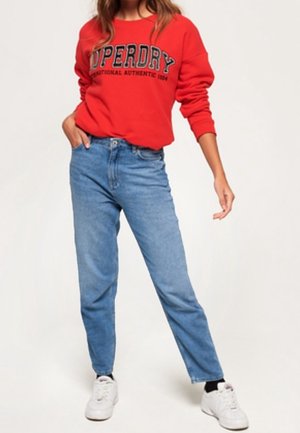 Vrouw draagt een rode Superdry trui, blauwe hooggetailleerde spijkerbroek en witte sneakers, staat met één hand in de zak.