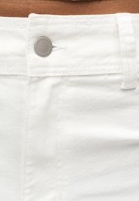 Λεπτομέρεια λευκού τζιν με ζώνη μέσης, μεταλλικό κουμπί, θηλιά ζώνης και ραφές στο ύφασμα.