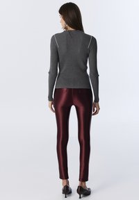 Woman wearing a ribbed gray long-sleeve top and shiny burgundy leggings, standing with back facing camera on a plain white background.