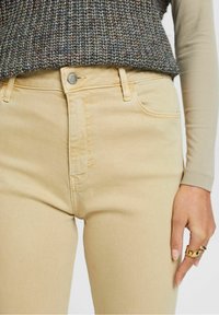 Jeans de talle alto beige con una textura suave, bolsillos frontales y cierre de botón metálico. Mano descansando sobre el muslo con un acento de anillo dorado.