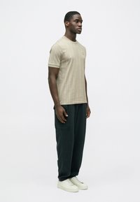 Camiseta beige de manga corta con patrón de texto vertical, combinada con pantalones de corte holgado en verde oscuro y zapatillas de color claro.