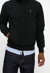 Veste légère noire avec col montant, poignets élastiques et ourlet. Présente un petit logo sur la poitrine et est superposée à une chemise bleu clair.