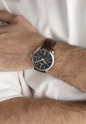 Ručni sat s crnim brojčanikom, oznakama od ružičastog zlata i smeđom kožnom narukvicom nošen na lijevoj ruci preko bijele tkanine.