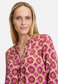 Blusa colorata con un motivo esagonale in tonalità di rosa, arancione e crema. Il tessuto ha una texture liscia con una leggera lucentezza.