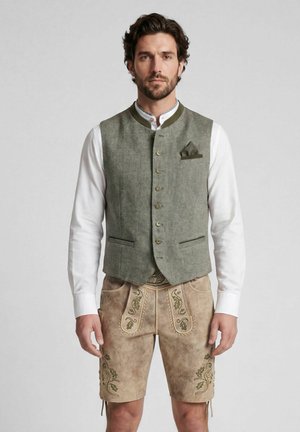 Man draagt een traditionele Beierse outfit met een groen vest, wit hemd met lange mouwen en beige geborduurde leren korte broek, rechtopstaand.