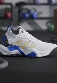 Scarpa sportiva bianca e blu con strisce beige appoggiata sul pavimento della palestra con kettlebell sfocati sullo sfondo.