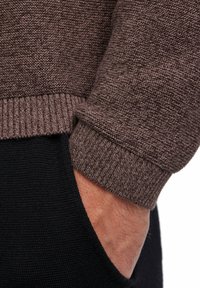 Brauner gestrickter Pullover mit gerippten Bündchen, kombiniert mit einem schwarzen strukturierten Kleidungsstück, das eine Hand in einer Tasche zeigt.