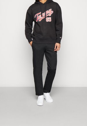 Man draagt een zwart hoodie met "Tommy 85" in rood en wit, zwarte broek en witte sneakers, staand tegen een effen lichte achtergrond.