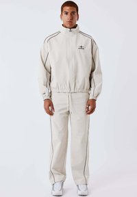 Man staat recht voor zich uitkijkend, gekleed in een licht beige trainingspak met zwarte biezen en witte sneakers, tegen een effen witte achtergrond.