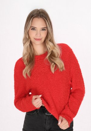 Pull rouge texturé à manches longues, col rond et coupe décontractée, associé à un pantalon noir. Détail de tissu clair et doux visible.