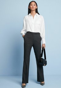 Weißes Button-Up-Hemd mit einer Tasche und langen Ärmeln, kombiniert mit dunkelgrauen geraden Hosen. Accessoire: kleine schwarze Handtasche.