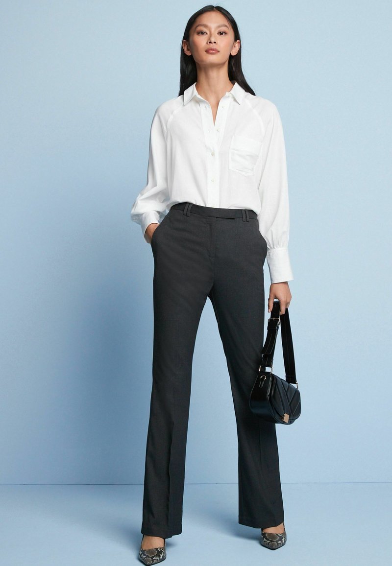 Weißes Button-Up-Hemd mit einer Tasche und langen Ärmeln, kombiniert mit dunkelgrauen geraden Hosen. Accessoire: kleine schwarze Handtasche.