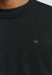 Un pull en tricot noir avec un col rond côtelé et un accent logo hexagonal en gris et jaune sur la poitrine.
