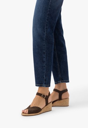 Pies con sandalias de cuña de ante marrón oscuro con tiras en el tobillo y hebillas doradas, combinadas con vaqueros de mezclilla azul oscuro.