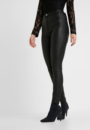 Nainen, jolla on mustat korkeavyötäröiset tekonahkahousut, musta pitsinen pitkähihaisen pusero ja mustat teräväkärkiset nilkkurit korkokengillä, seisoo valkoisella lattialla.