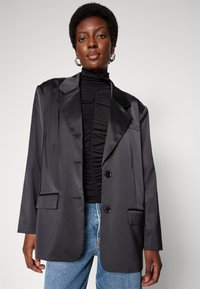 Blazer en satin noir avec revers cranté, deux poches avant et fermeture à double bouton, associé à un col roulé texturé noir et un jean.