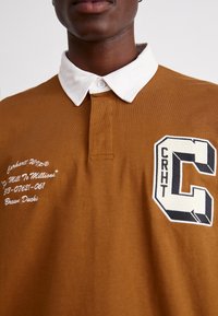 Brun polo-skjorta med vit krage; har broderad logotyp "CRHT" och text. Slät tyg, korta ärmar och knapplåsning vid halsen.