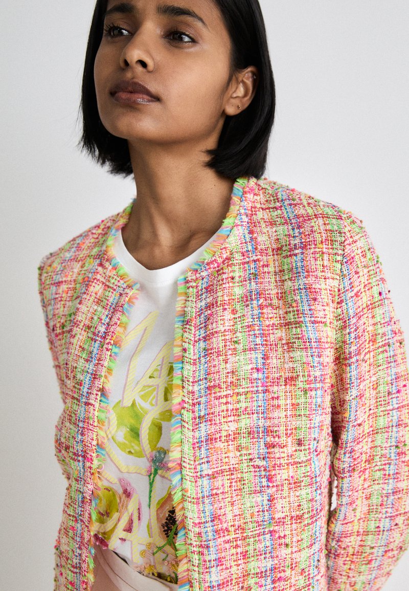 Mehrfarbige Tweedjacke mit einem strukturierten, ausgefransten Rand, die pinke, grüne und blaue Fäden aufweist, getragen über einem weißen T-Shirt mit Grafik.