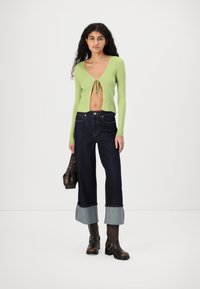 Top de manga larga de rayas verdes con un lazo en la parte delantera, jeans de mezclilla oscuros de cintura alta con dobladillos remangados, y botas marrones hasta el tobillo con hebillas.