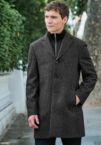 Grå herringbone ullkappa med slagkrage, knäppning fram och två sidofickor; bärs över en svart zip-up jacka.