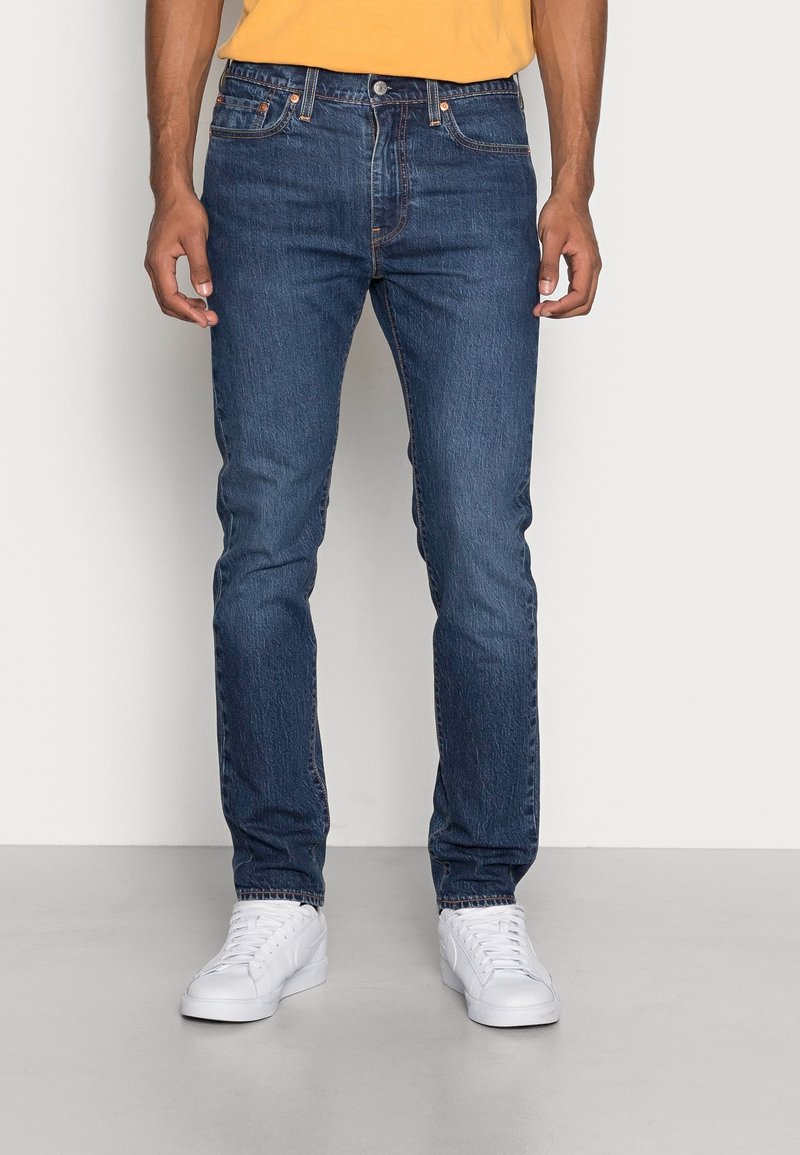 Hombre vistiendo jeans azul ajustados, zapatillas blancas y una camiseta amarilla, de pie contra un fondo claro y liso.