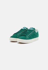 Sneakers de camurça verde com uma sola de borracha branca, apresentando ilhós de metal, atacadores verdes e um emblema azul na língua.