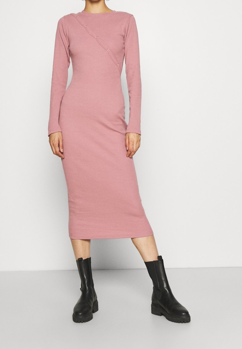 Långärmad, ribbad klänning i ljusrosa med en figursydd silhuett, som har en sned söm och dekorativa knappar på ena sidan. Matchas med svarta ankla stövlar.