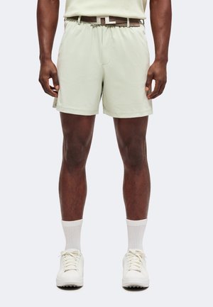 Mann trägt hellbeige Shorts, weiße Kniesocken, weiße Turnschuhe und einen braunen Gürtel, steht vor einem einfarbigen Hintergrund.