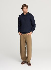 Maglione a trecce in maglia blu navy con scollo rotondo, abbinato a pantaloni kaki e scarpe marroni. Il modello è in piedi con le mani nelle tasche.