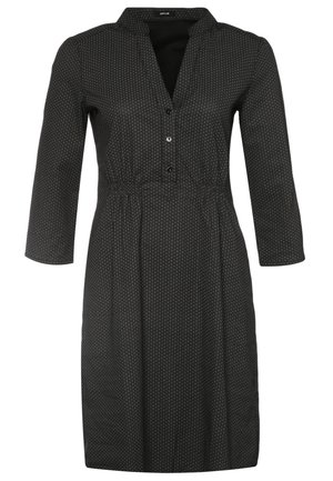 Femme portant une robe noire à motifs, manches longues, longueur genou, des collants noirs, des bottines et tenant une pochette noire.