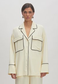 Crème overhemd met knopen en zwarte piping, voorzien van twee voorzakken en een losse fit. Gemaakt van een soepele stof met een kraag en lange mouwen.