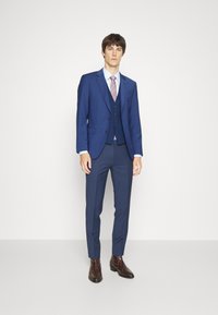 Costum albastru cu sacou ajustat și vestă cu trei butoane. Combinație cu cămașă bleu deschis și cravată cu model. Botine maro texturate.