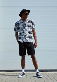 Short-sleeved black and white floral print shirt paired with black shorts and black sneakers, completed with a black wide-brimmed hat and sunglasses.