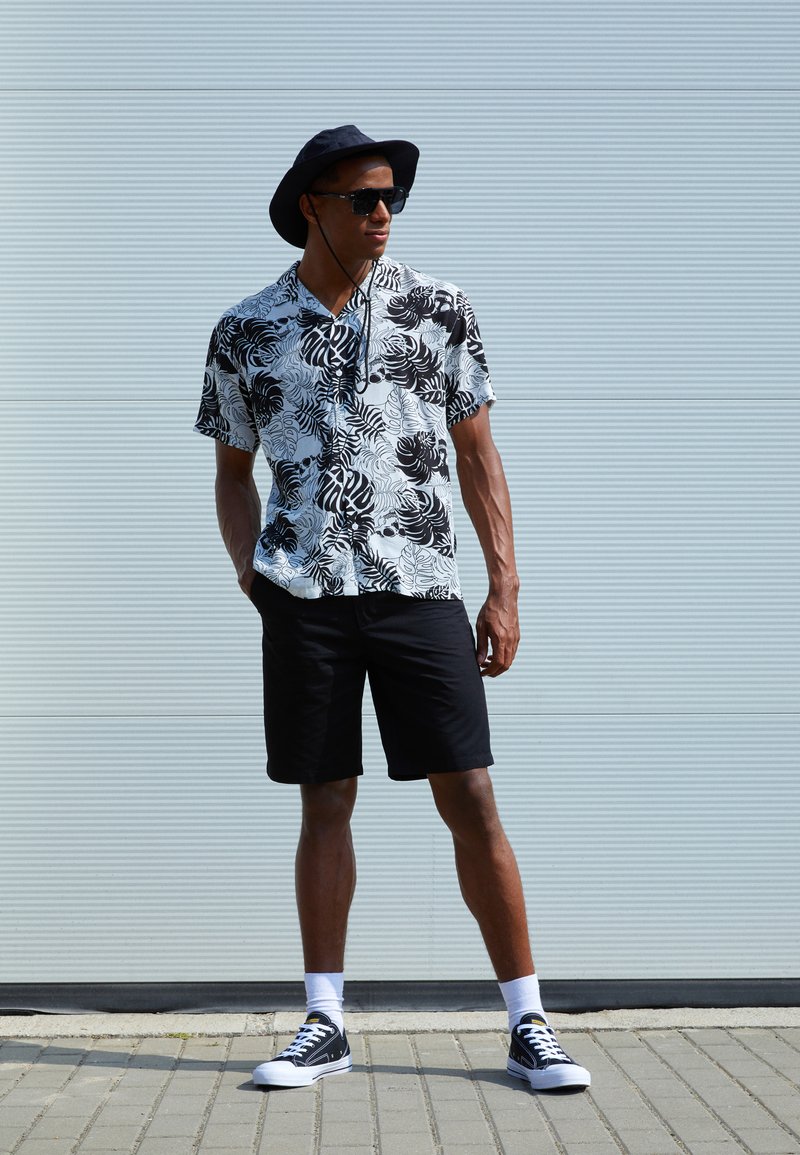 Short-sleeved black and white floral print shirt paired with black shorts and black sneakers, completed with a black wide-brimmed hat and sunglasses.
