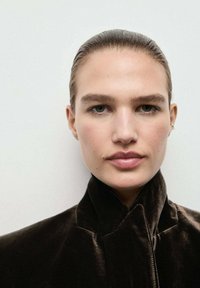 Joven con el cabello peinado hacia atrás que lleva una chaqueta de terciopelo oscuro con cuello alto, mirando de frente contra un fondo claro y liso.