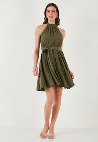 Abito halter verde oliva con busto aderente, gonna a strati e cintura con un nastro. Presenta una texture liscia e una silhouette fluttuante.
