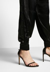 Svarta satinjoggers har sidoknappar och elastiska cuffs. Matchas med svarta högklackade sandaler med remmar, som visar en minimalistisk design.