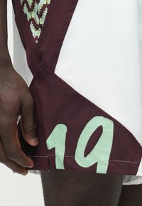 Tumši sarkanas un baltas sporta šorti ar teksturētu paneli, sānos ar mint zaļas krāsas ciparu "19" un sarežģītiem rakstiem. Audums ir viegls.