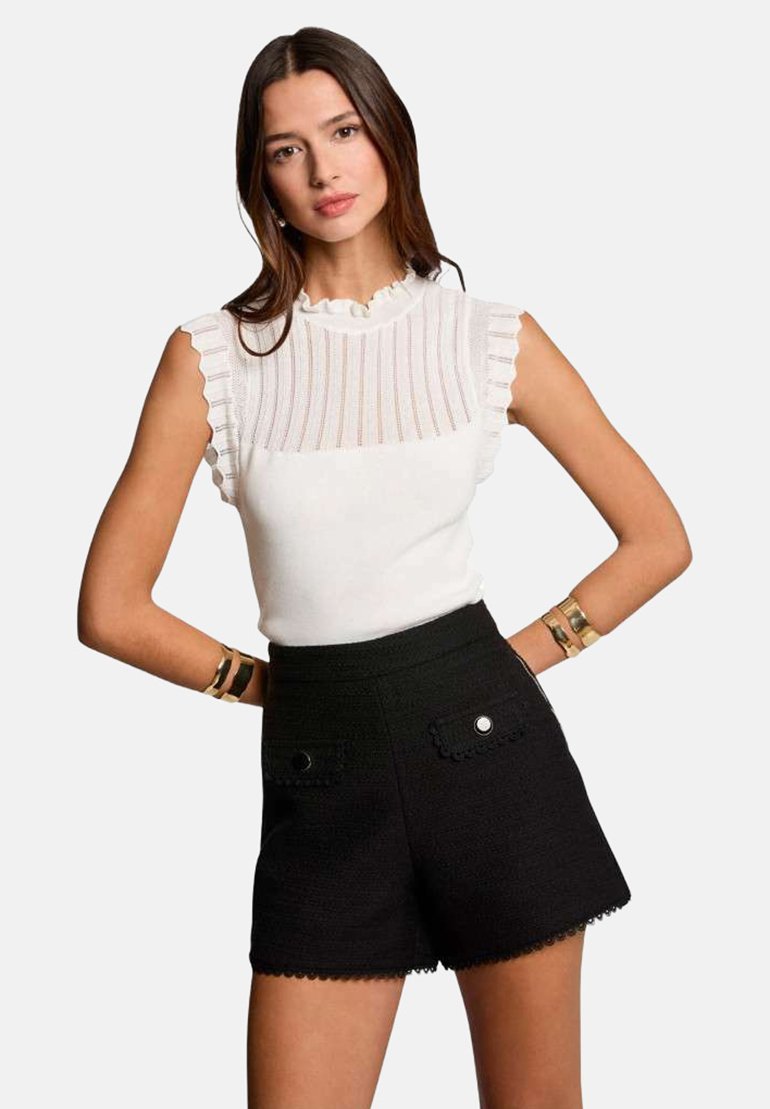 Haut en tricot blanc à côtes avec des manches volantes, associé à un short noir taille haute avec des bords festonnés et des boutons décoratifs.